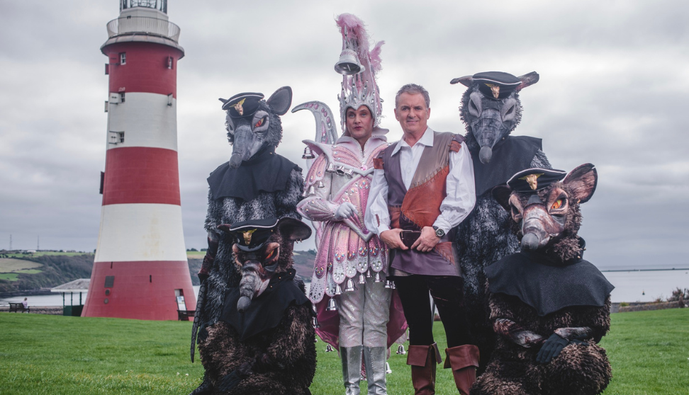 A group of people in theatrical costumes pose on a grassy area in front of Plymouth Hoe’s iconic red and white lighthouse. Four wear detailed rat costumes with naval-style hats and black capes, while another person is dressed in a sparkling, feathered outfit with silver boots. One other wears a pirate-style shirt and boots. The sky is cloudy and the sea is visible in the background.